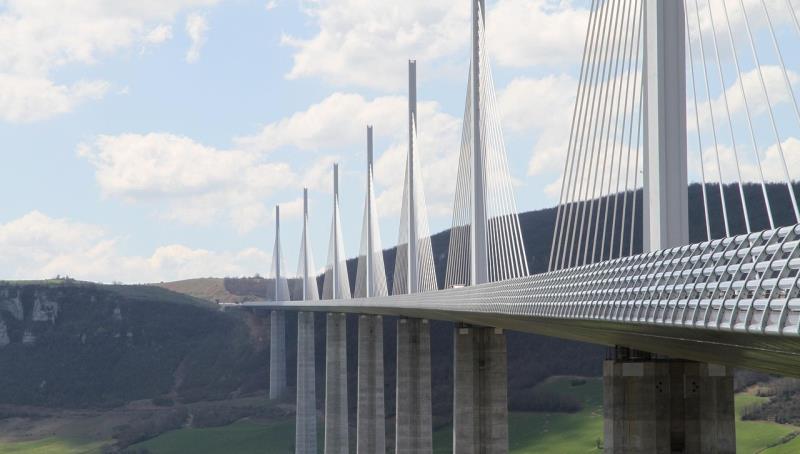 A large cable-stayed bridge with tall concrete pylons and steel cables, set against green hills and a blue sky.