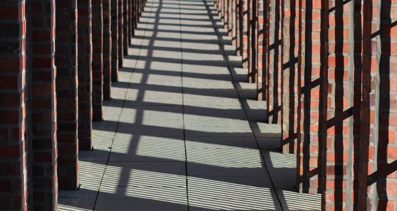 Photograph of a long corridor with brick columns casting striped shadows on a tiled floor. The alternating light and shadow create a repeating geometric pattern emphasizing depth and perspective.