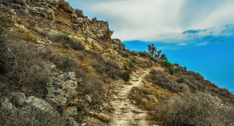 There's a narrow, sandy path going up a mountain or hillside. The ground is mainly brown and covered in rocks and scrubby bushes. Over the hill a corner of blue sky can be seen. 